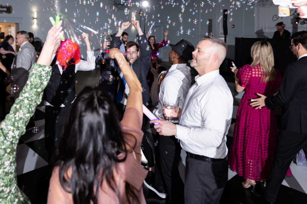 A packed dance floor filled with wedding guests celebrating under floating bubbles inside a modern wedding venue in North Georgia. Friends raise their arms, hold glow sticks, and dance closely together beneath crystal chandeliers and soft wall lighting, creating a high-energy reception scene.