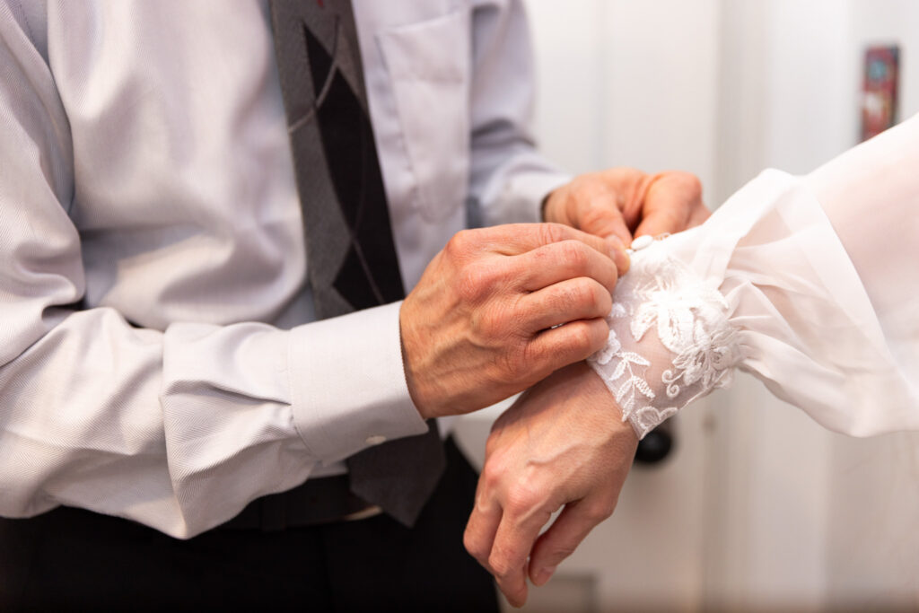 Close-up of the groom fastening the delicate lace cuff of the bride’s sleeve, carefully buttoning the sheer embroidered fabric as part of their shared getting-ready process for their living room elopement. His gray shirt and patterned tie contrast against the intricate white lace detail.
