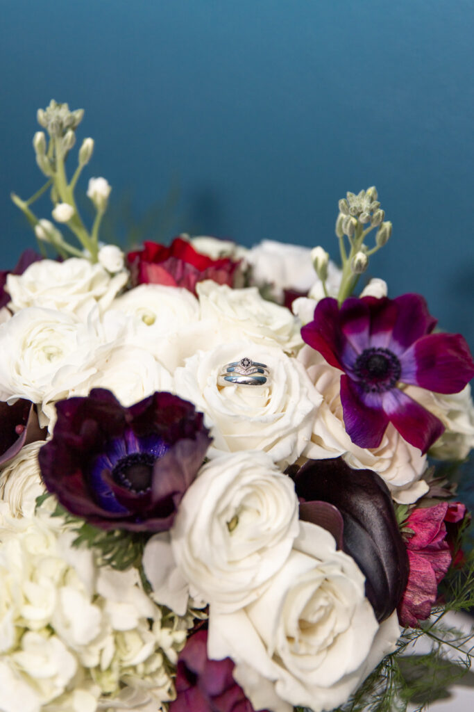 Close-up of the bride’s bouquet arranged with white roses, hydrangeas, and deep purple flowers, with her wedding rings resting on a bloom at the center — a floral detail photographed as part of their living room elopement celebration.