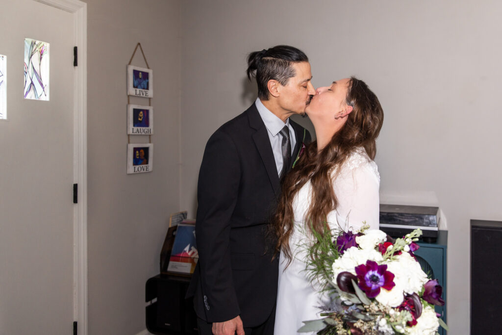 The couple share their first kiss as husband and wife, standing close together in their living room. The bride tilts her face upward, bouquet cascading with white hydrangeas and dark purple blooms, while the groom leans in gently. A small wall hanging that reads “Live, Laugh, Love” appears behind them.