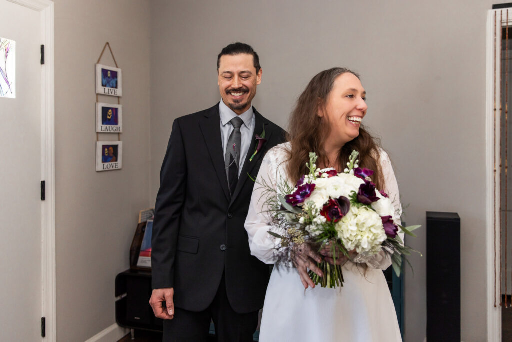 The bride laughs brightly, turning slightly away from the groom as he smiles behind her. She holds her bouquet in both hands, silver flats visible beneath her knee-length ivory dress. The relaxed energy of the moment feels spontaneous and joyful.