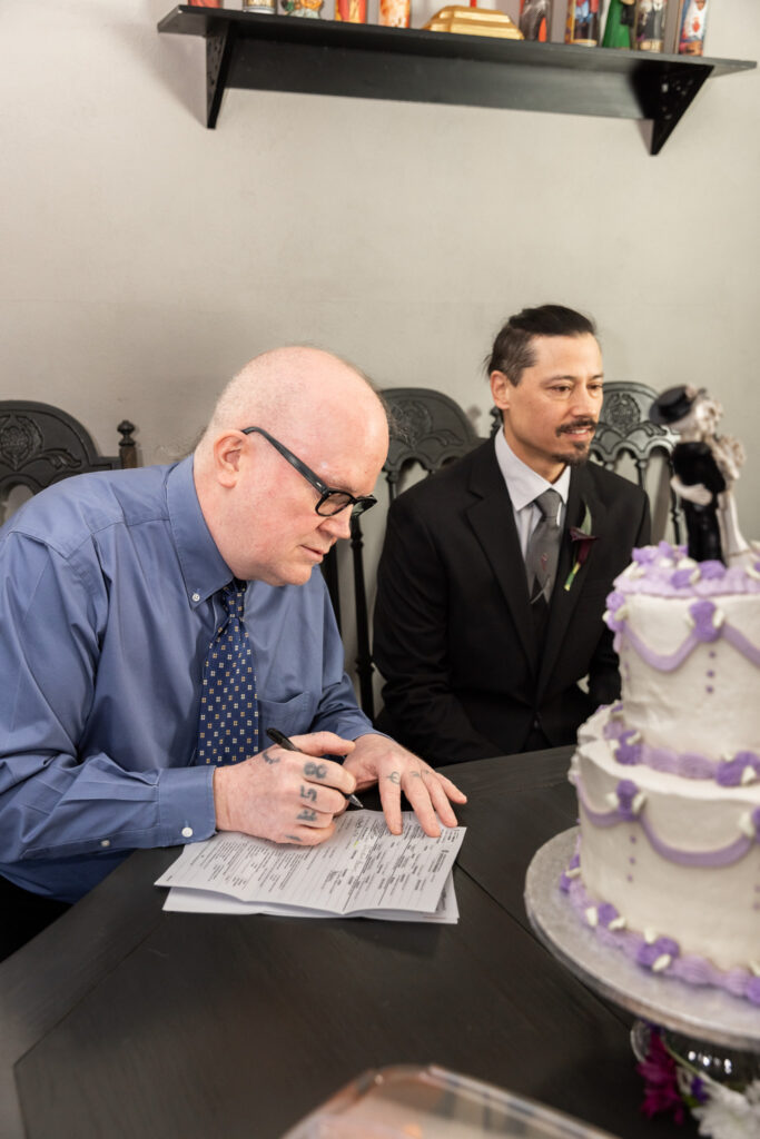 The officiant signs the marriage license at the dining room table, pen in hand, concentrating on the paperwork. A two-tier white cake with lavender piping and a skeleton bride and groom topper sits prominently in the foreground.