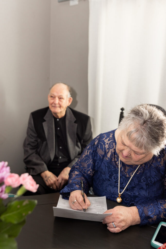 A woman in a navy lace dress signs the marriage license as a man seated behind her watches with a gentle smile. Soft light falls across the table, with pink flowers slightly blurred in the foreground.