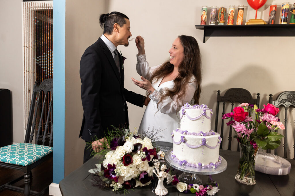 The bride feeds the groom a bite of cake while laughing, her hand lifted toward his mouth. He leans in playfully, smiling, as the cake and bright pink flowers sit on the table between them. The mood is lighthearted and affectionate.