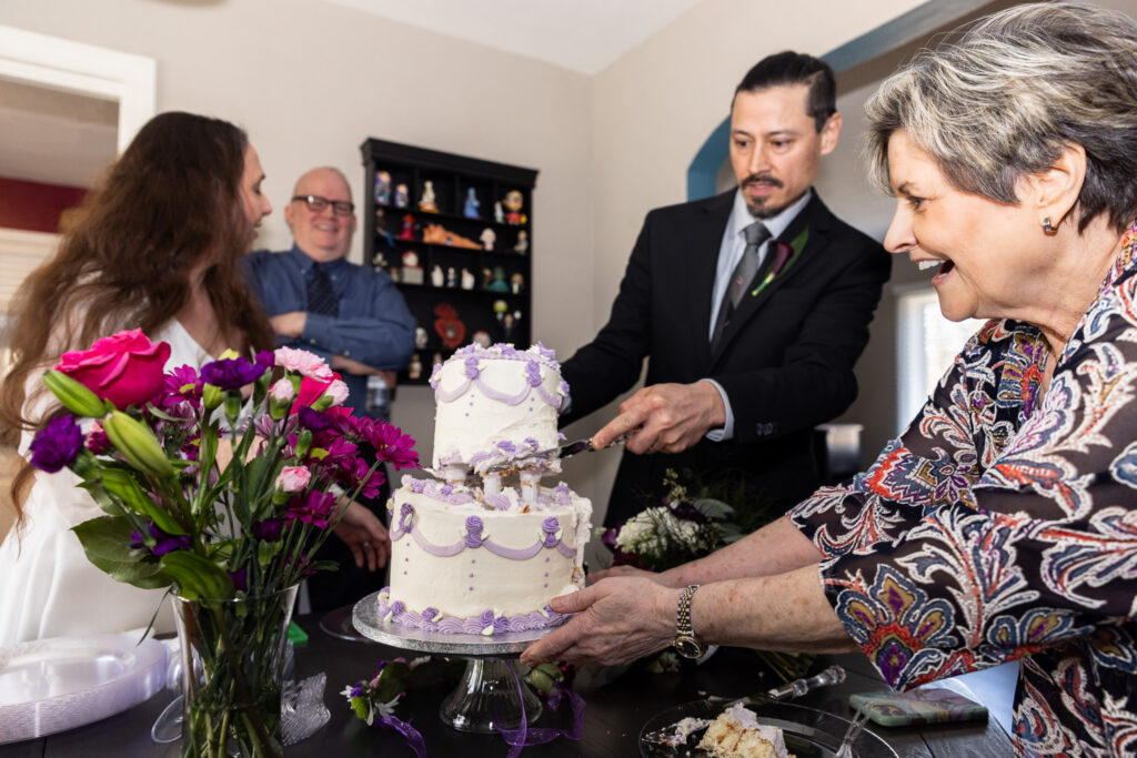 The groom cuts into the two-tier lavender-trimmed cake while a woman in a patterned jacket steadies the cake stand with both hands. The bride stands nearby laughing, and the officiant smiles in the background. A vase of bright pink and purple flowers fills the foreground, adding color to the lively moment.