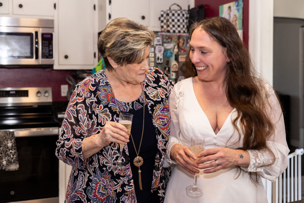 The bride and her mother in law stand close together in the kitchen, each holding a champagne flute. They smile warmly at one another, sharing a quiet celebratory moment after the ceremony.
