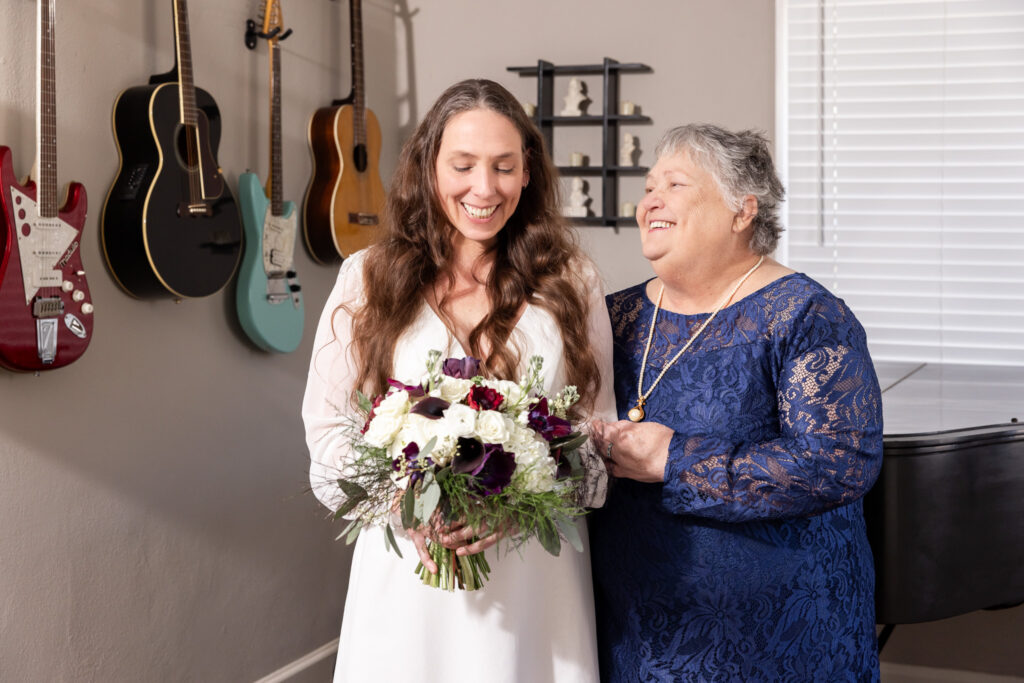 The bride stands beside her mom in a navy lace dress, both smiling as they pose together. The bride holds her bouquet low, and several guitars and a piano are visible behind them.