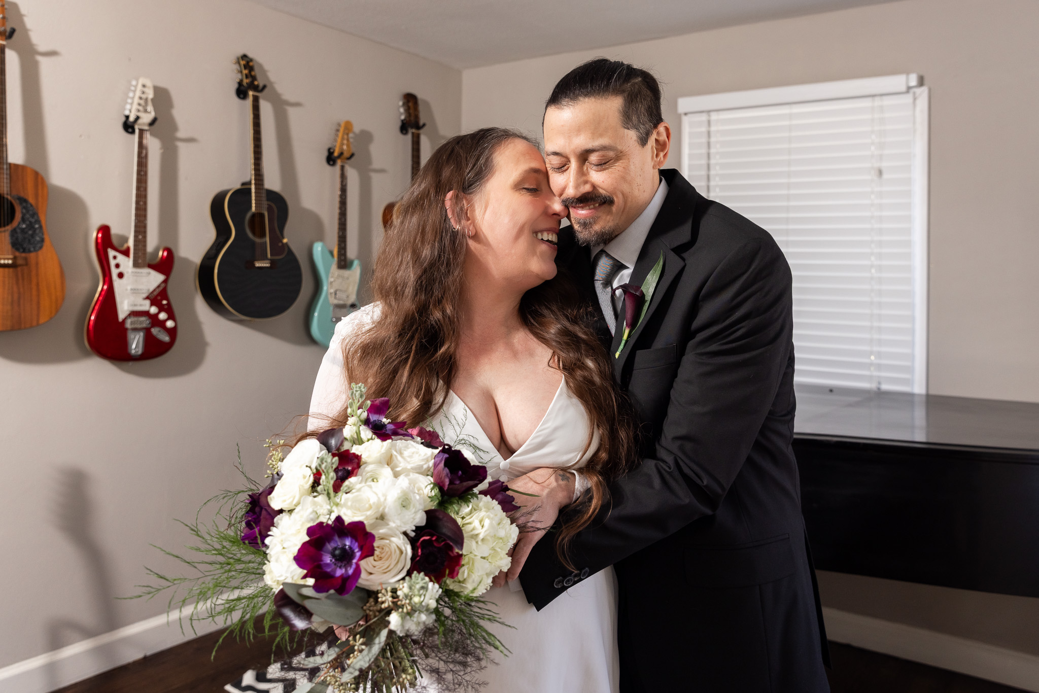 The bride and groom stand wrapped in each other’s arms in front of a black baby grand piano, guitars mounted along the wall behind them. She holds a bouquet of white hydrangeas, roses, and deep burgundy blooms while smiling up at him, and he leans his forehead against hers during their living room elopement portraits.