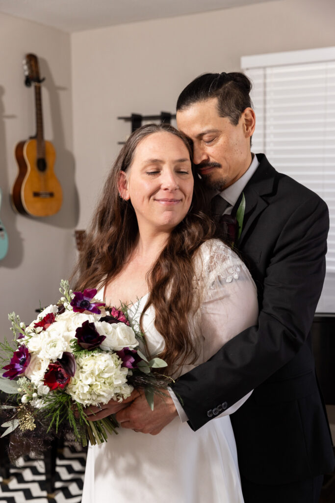 The bride closes her eyes and smiles softly as the groom embraces her from behind, both standing in front of the baby grand piano during their living room elopement. Her bouquet rests in her hands while his arms wrap securely around her waist, guitars hanging neatly in the background.