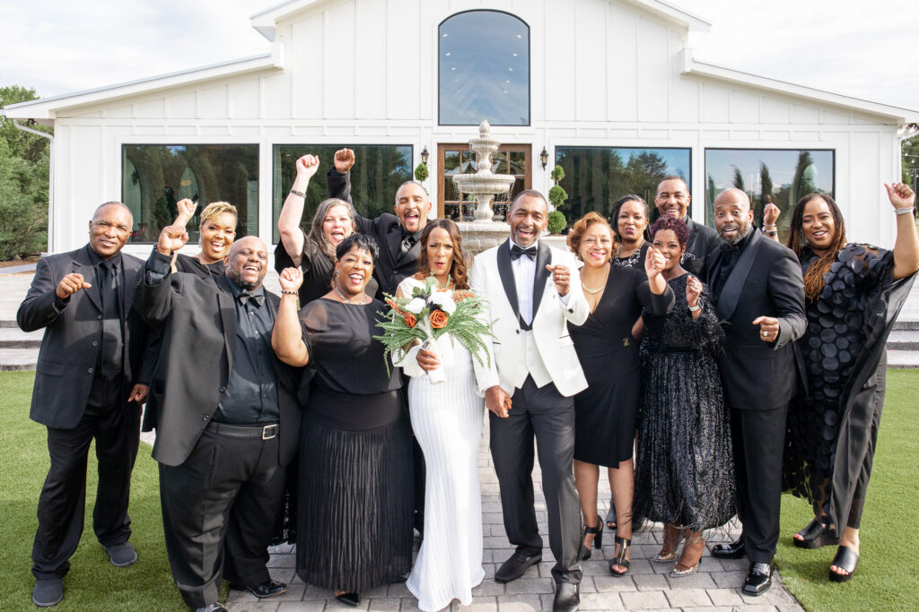 The bride and groom stand at the center of a large group of joyful family members and friends gathered outside a bright white wedding venue. The couple stands arm in arm in the front row, the bride holding a bouquet of orange roses and greenery while guests around them cheer with raised fists and wide smiles. Everyone is dressed in elegant black formalwear, creating a striking visual contrast against the white building and stone fountain behind them. The group stands on a paved walkway bordered by green lawn, with the venue’s tall windows and symmetrical architecture framing the celebratory moment.