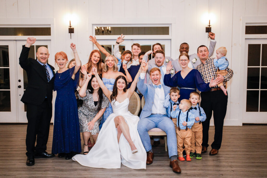 The bride and groom sit together on chairs in the center of the dance floor while their friends and family stand behind them cheering with raised arms during an energetic photo dash moment at the wedding reception.