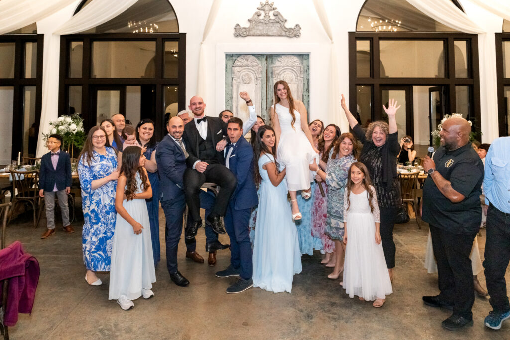 Guests lift the bride and groom into the air while laughing and celebrating during a playful photo dash group photo on the reception floor surrounded by elegant arches and candlelight.