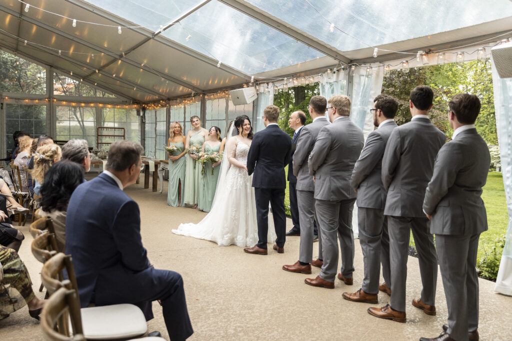 A couple stands at the front of a clear-top tent during their ceremony, surrounded by their wedding party, showcasing a rainy wedding day plan that still allows natural light to filter through. String lights and greenery decorate the space while guests sit along the sides under the covered structure.
