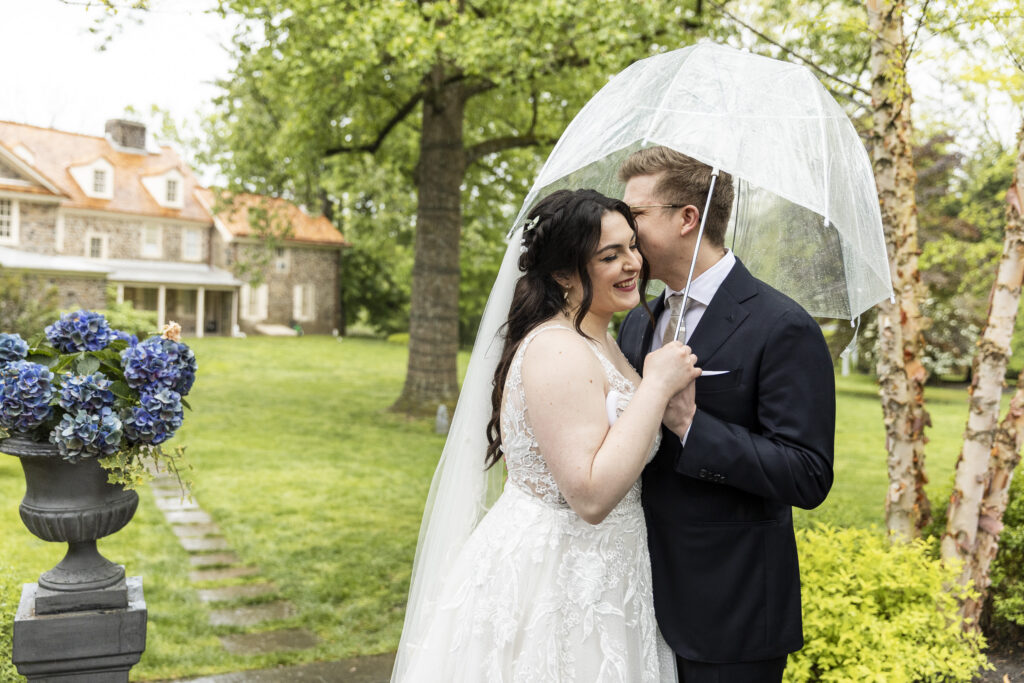 A bride and groom stand close together under a clear umbrella in a lush garden, smiling softly as the groom leans in toward her cheek. The bride’s lace gown and veil fall gently over her shoulders, while the groom wears a dark suit. Behind them, a stone house and greenery create a calm, romantic backdrop with soft, diffused light from the rain.
