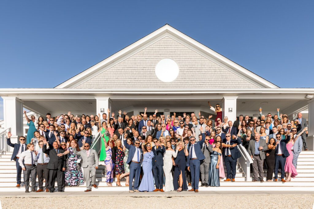 The bride and groom stand centered on the steps outside a coastal-style venue while dozens of guests wave and celebrate during a joyful big group photo in bright afternoon sunlight.