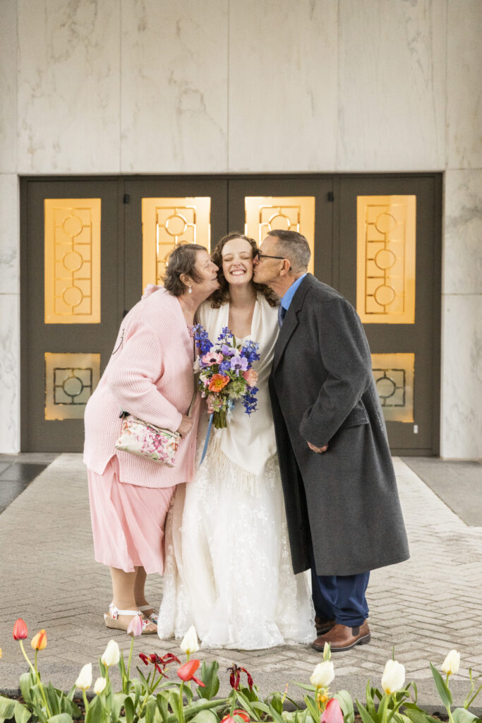 A bride stands between two older family members who kiss her cheeks simultaneously, her eyes closed and smiling while holding a colorful bouquet, a joyful backup plan moment captured under a covered entryway with soft glowing doors behind them and tulips in the foreground.