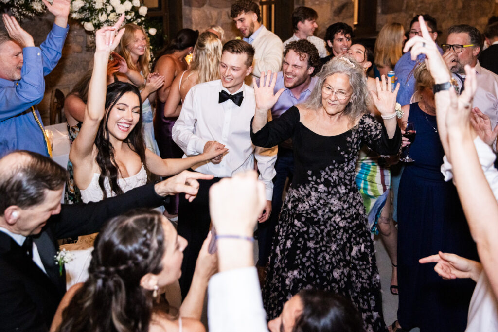 A packed dance floor filled with wedding guests moving and dancing together under warm reception lighting. At the center of the frame, an older woman with gray hair lifts both hands joyfully while dancing beside younger guests who clap and raise their arms in rhythm to the music. People surrounding them laugh, sing along, and point playfully toward one another as the energy of the dance floor builds. The background shows guests crowded closely together, creating a lively atmosphere of celebration and shared excitement.