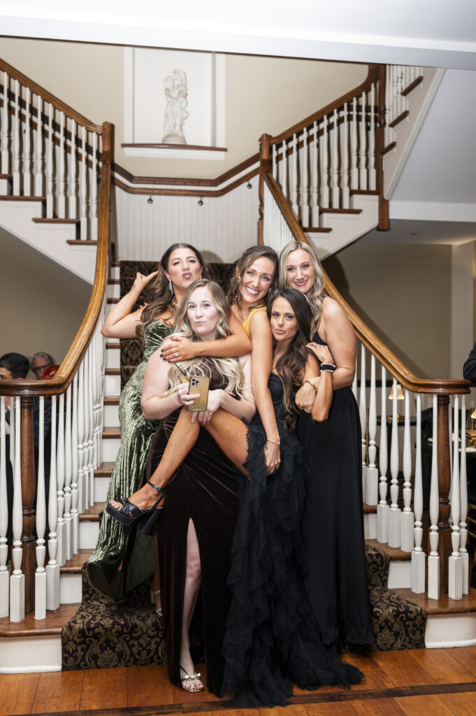 A group of wedding guests pose together on a grand staircase inside a reception venue, laughing and leaning into each other. They wear a mix of formal dresses in black, green, and neutral tones. The staircase features white railings and warm wood accents, creating a classic, elegant indoor setting.