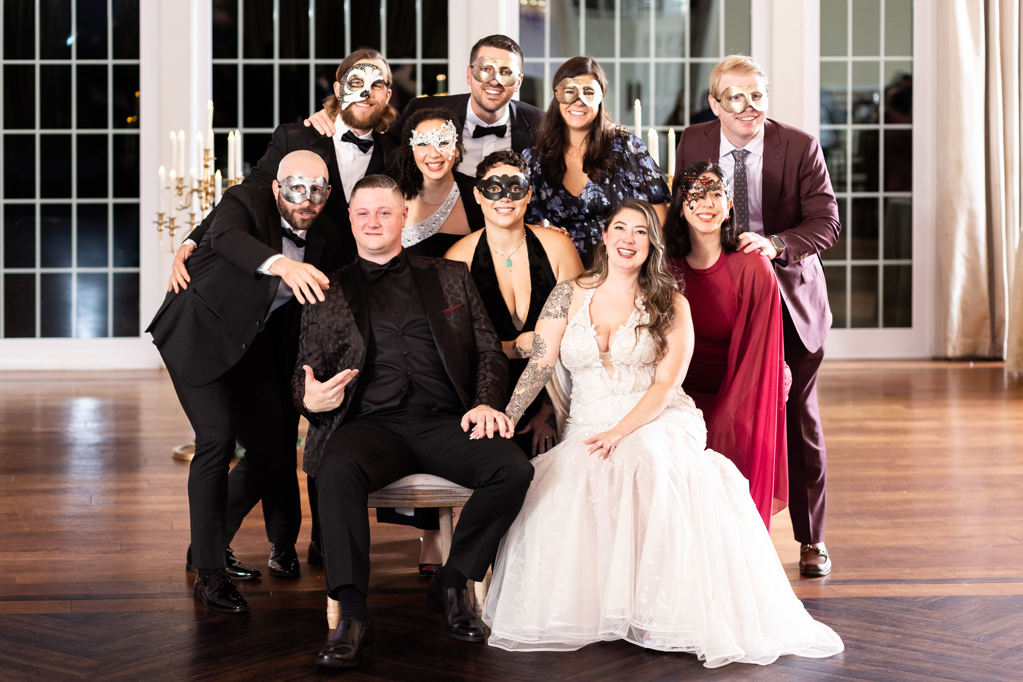 Friends gather closely behind the bride and groom wearing masquerade masks while pointing and laughing during a playful photo dash photo on the reception dance floor.