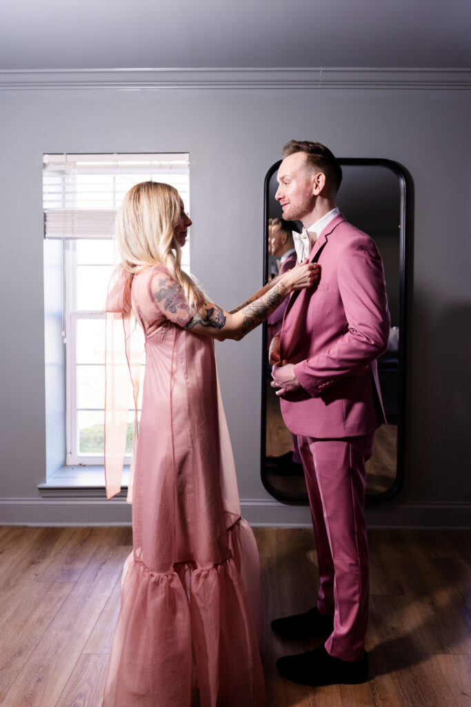 In a softly lit getting-ready room at a wedding venue in North Georgia, a woman in a flowing blush dress gently adjusts the boutonniere on a groom wearing a tailored pink suit and cream bow tie. Natural light streams through a nearby window, highlighting the intimate pre-ceremony moment.