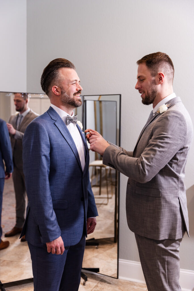 Inside a bright preparation suite at a wedding venue in North Georgia, one groom in a steel-blue suit smiles warmly as another man in a light gray suit carefully pins a white boutonniere to his lapel. A full-length mirror behind them reflects the moment, capturing the quiet anticipation before the ceremony.