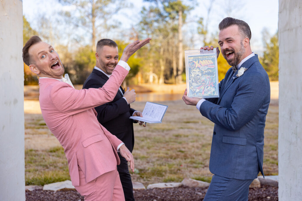 The grooms pose playfully during their ceremony, one dramatically presenting a framed vintage comic book while the other gestures toward it with an exaggerated flourish. The officiant stands behind them holding a ceremony notebook. The outdoor setting features soft winter grass and trees in the background, highlighting the couple’s personality and joy during their same-sex wedding ceremony.