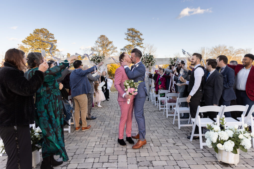 The newly married grooms share a kiss in the center of the aisle as guests on both sides wave ribbon wands overhead in celebration. The groom in pink holds a bouquet of blush ranunculus, ivory roses, and greenery, while the groom in blue wraps an arm around him. White ceremony chairs line the cobblestone aisle, and floral arrangements anchor the space. An lgbt wedding photographer captures the joy and movement of this triumphant recessional moment.