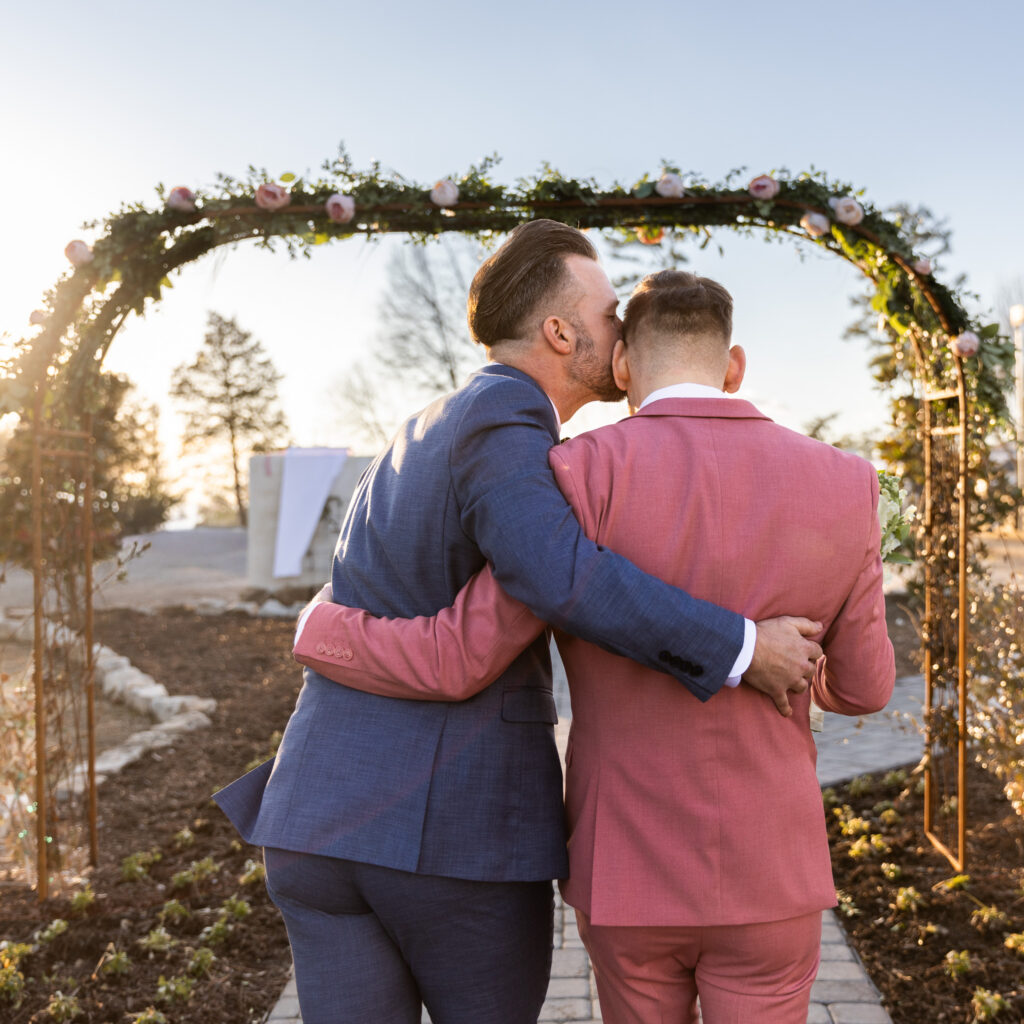 From behind, the two grooms walk arm in arm down a stone pathway beneath a circular greenery arch decorated with blush roses. Golden sunlight outlines their blue and pink suits as one groom kisses the other’s temple. This warm, backlit portrait showcases the romantic storytelling style central to lgbt wedding photography.