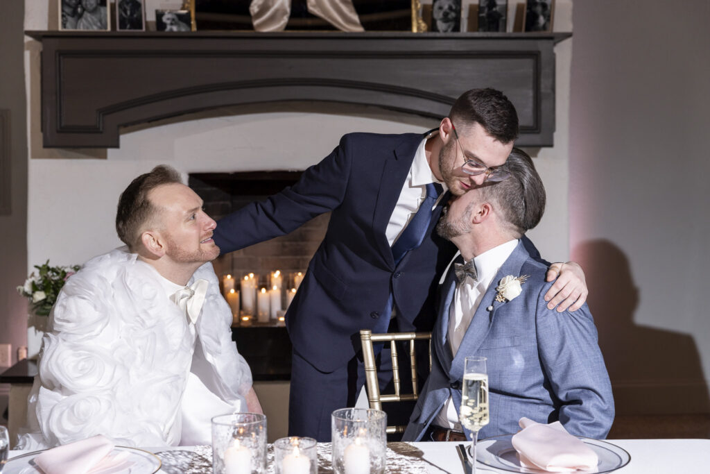 Three men are together at a sweetheart table during the reception, sharing an affectionate moment. Two grooms sit side by side in elegant suits while a friend leans down between them to kiss one of them on the cheek. The table in front of them is decorated with glowing candles, champagne flutes, and neatly folded blush-colored napkins. Behind them, a fireplace mantel lined with framed photos and candles creates a cozy, intimate atmosphere with soft, warm lighting.