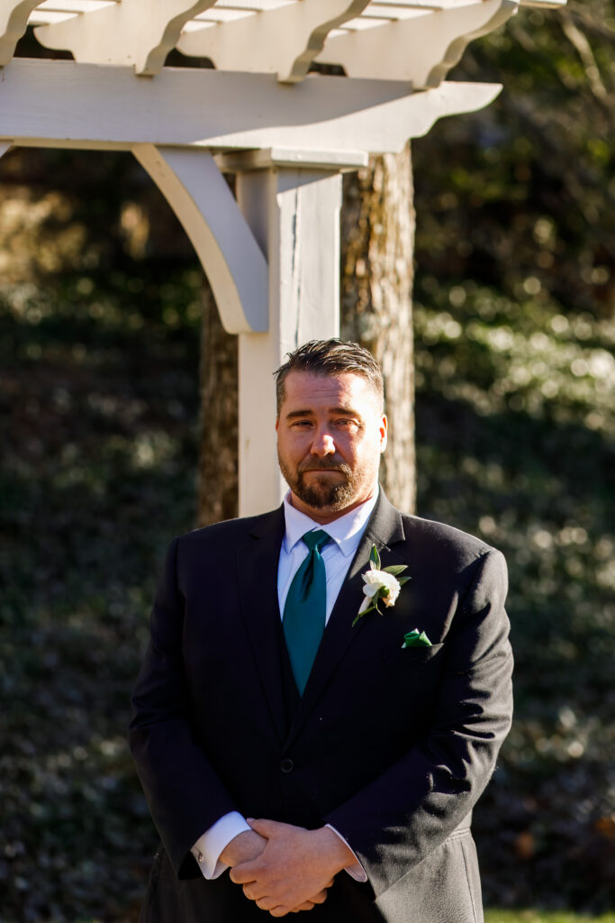 At a Philadelphia wedding ceremony, the groom stands alone beneath a white wooden arbor, his hands gently clasped in front of him. He wears a black suit with a deep green tie and a white rose boutonniere pinned to his lapel. His expression is steady and slightly emotional, as warm afternoon sunlight highlights one side of his face while the background fades into soft greenery.