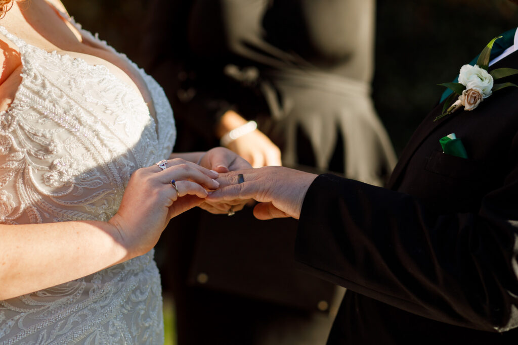A close, intimate moment during a Philadelphia wedding ceremony shows the bride placing a ring onto the groom’s finger. The bride’s lace dress and engagement ring are visible as her hands gently guide the band into place. The groom’s hand is steady, his black suit sleeve and boutonniere partially visible. Warm golden light falls across their hands, emphasizing the emotion and significance of the exchange.