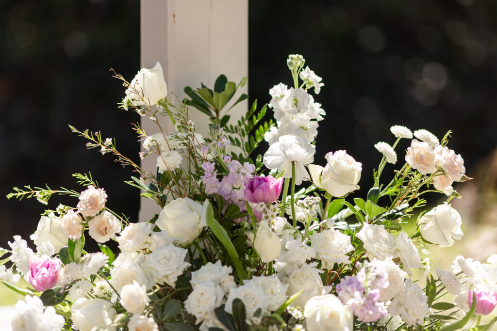A close-up of a Philadelphia wedding floral arrangement shows a lush mix of white roses, pale blush flowers, and delicate lavender blooms, interwoven with greenery. Sunlight illuminates the petals, creating bright highlights and soft shadows, while the background falls into a dark blur, making the flowers appear vivid and three-dimensional.