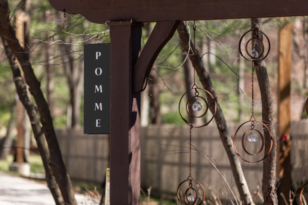 A wooden post at a Pomme Radnor wedding displays a vertical black sign reading “POMME” in white letters. Hanging beside it are delicate metal ornaments made of circular rings with small glass crystals suspended in the center, gently catching the light. The background shows a wooded setting with bare tree branches and a soft, natural landscape.