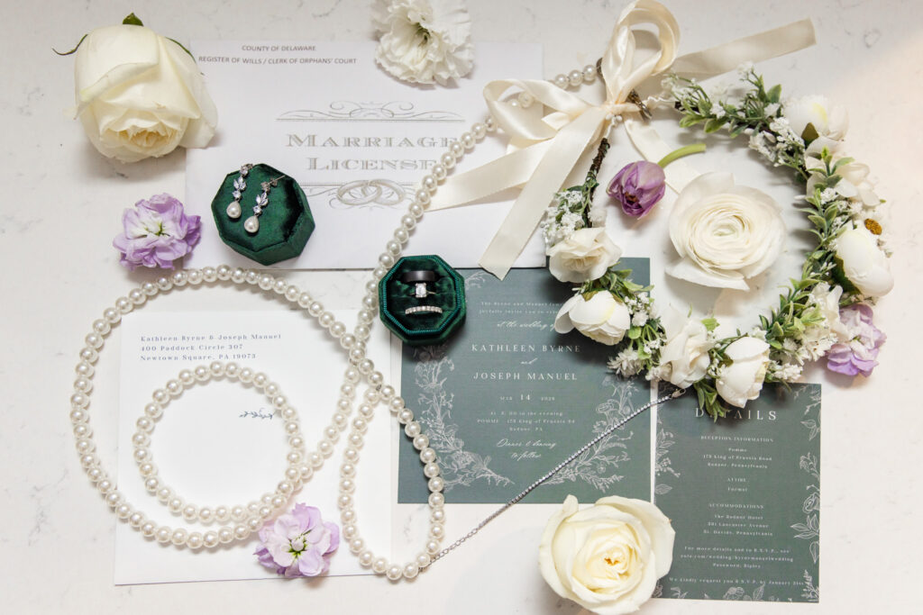 A flat lay of wedding details arranged on a light marble surface, including a pearl necklace draped in soft loops, a pair of pearl drop earrings in a green velvet box, wedding bands in a matching ring box, and floral accents in white and lavender tones. Invitation cards with elegant typography are layered beneath the accessories, creating a cohesive and refined composition.