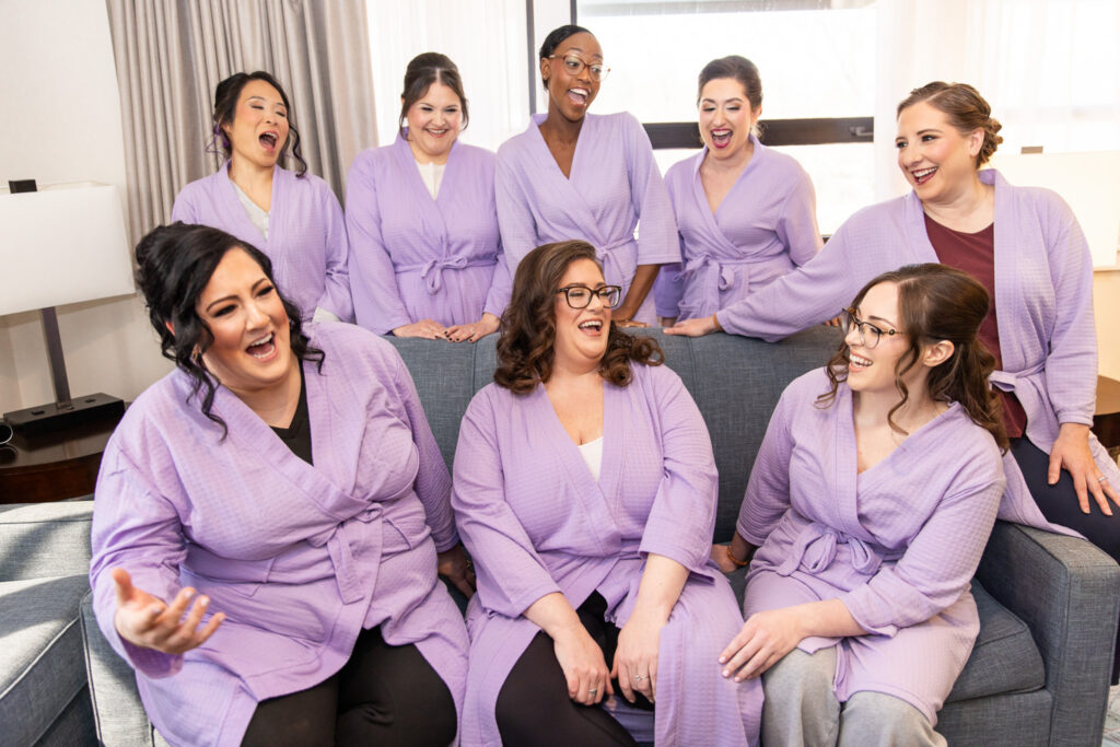 A group of bridesmaids in matching lavender robes gather in a bright getting-ready space, sitting and standing around a couch. They laugh and react animatedly to something off-camera, their expressions joyful and full of anticipation. Soft window light illuminates their faces, highlighting the excitement and camaraderie of the morning.