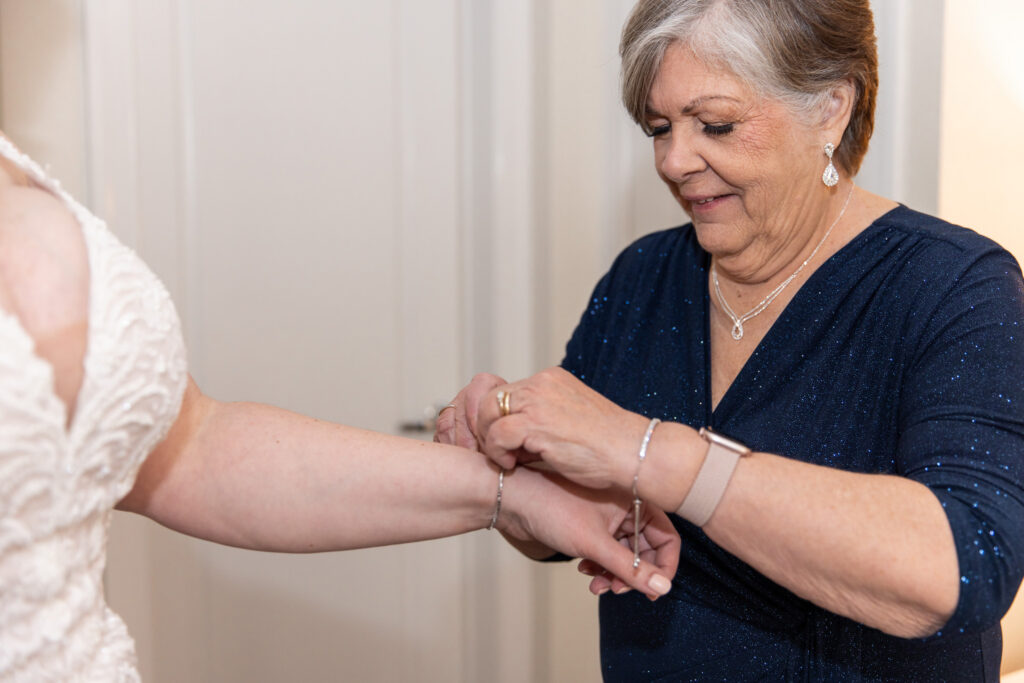 During Philadelphia wedding preparations, an older woman with short gray hair and a navy, subtly sparkling dress carefully fastens a delicate bracelet around the bride’s wrist. Only part of the bride is visible—her arm and the side of her lace gown—while the woman focuses intently, her expression warm and concentrated. Soft indoor lighting and neutral walls create a calm, intimate setting.