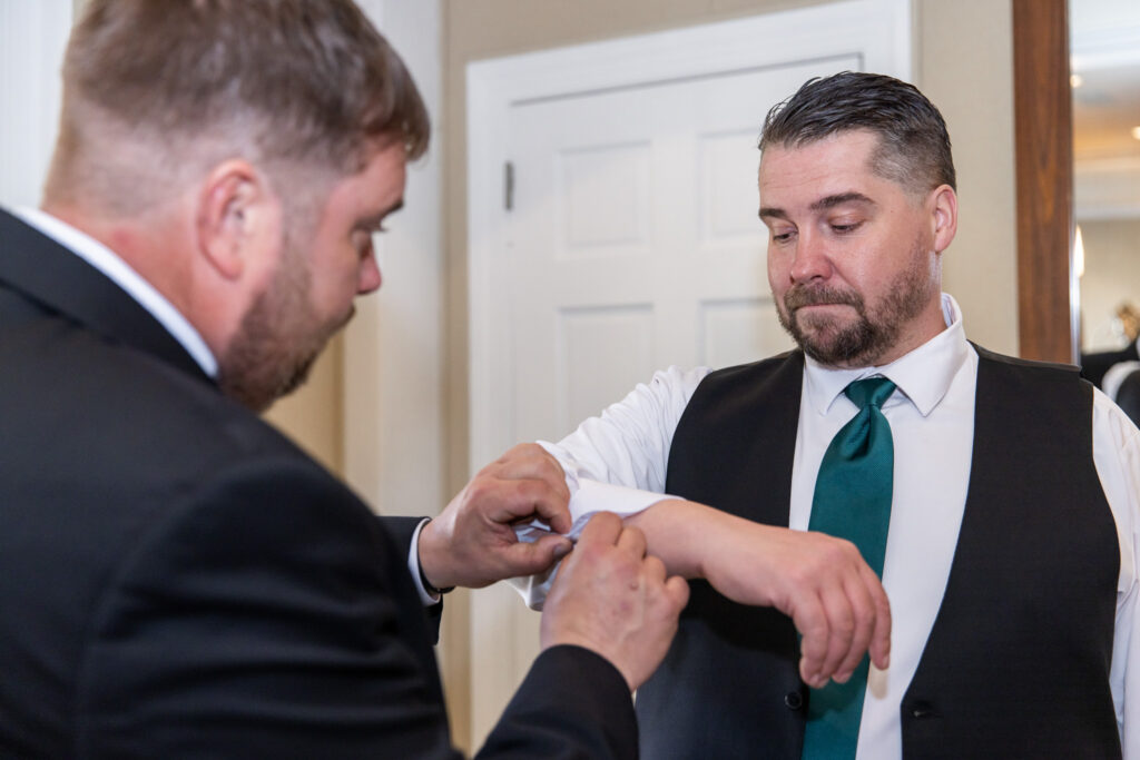 In a close-up from Philadelphia wedding preparations, one man adjusts another man’s shirt cuff, carefully fastening a cufflink. The groom or groomsman wears a white dress shirt, dark vest, and a deep green tie, while the helper’s hands are centered in the frame. The focus on the hands and fabric highlights the quiet, detailed moments before the ceremony.