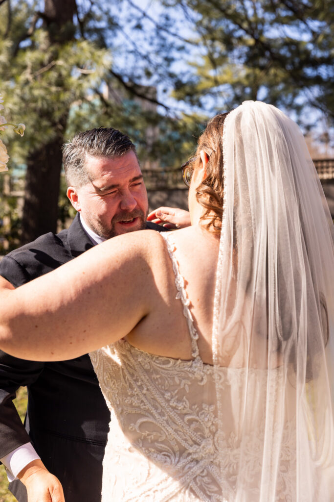 A close, intimate moment during a Pomme Radnor wedding shows the bride and groom embracing outdoors. The bride’s back faces the camera, revealing the detailed lace of her gown and a long veil cascading down her back. The groom’s face is partially visible over her shoulder, his eyes slightly closed as he begins to cry during their first look, as he leans into the hug, surrounded by soft greenery and warm sunlight.