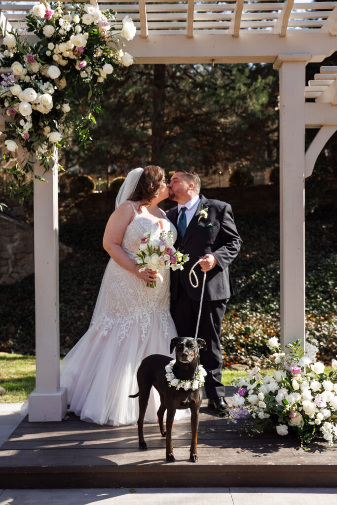 At a Pomme Radnor wedding, the bride and groom share a kiss beneath a white arbor decorated with lush flowers. The bride holds her bouquet close, while the groom gently leans in. In the foreground, a black dog wearing a floral wreath stands alert on a leash, centered in the frame, adding a charming and personal touch to the moment.