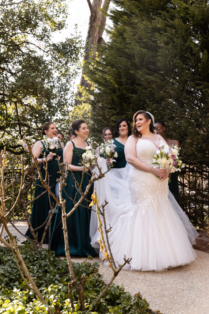 The bride walks along a garden path with her bridesmaids gathered behind her, all holding bouquets. The bridesmaids wear matching deep green dresses, while the bride’s gown flows softly as she moves. Sunlight filters through tall trees and greenery, casting dappled light across the group as they laugh and interact naturally.
