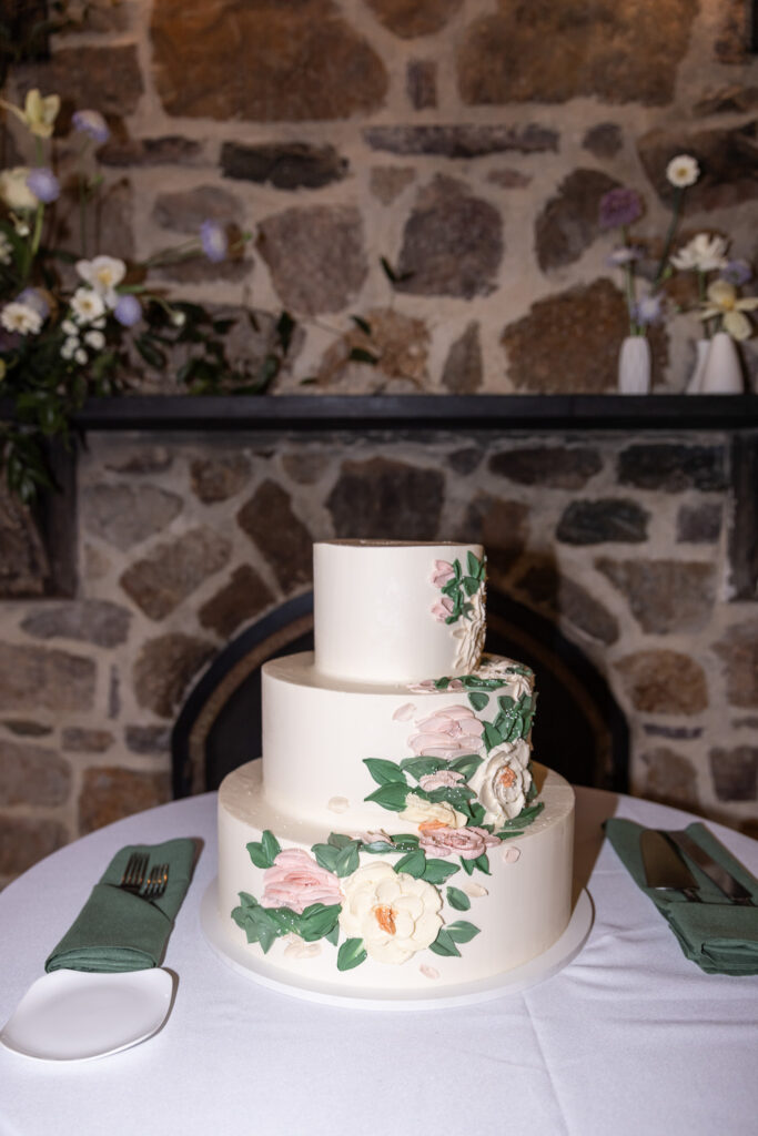 A three-tiered wedding cake sits centered on a round table at a Pomme Radnor wedding reception. The cake is decorated with soft, hand-piped flowers in blush, ivory, and green tones that cascade diagonally down the tiers. On either side of the cake, green cloth napkins are neatly folded beside silverware, with a rustic stone fireplace forming the textured backdrop.
