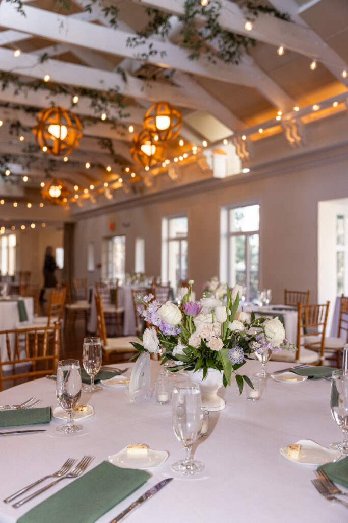 A close-up view of a reception table at a Pomme Radnor wedding shows a floral centerpiece filled with white roses, soft pink blooms, and greenery. Surrounding the arrangement are clear water glasses, folded green napkins, silverware, and small plates with butter. In the background, the softly blurred room reveals more tables and glowing string lights above.