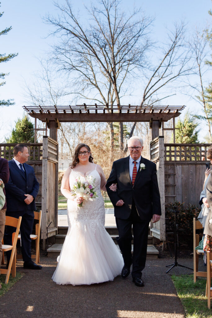 At a Pomme Radnor wedding ceremony, the bride walks down the aisle arm-in-arm with her father. She holds a bouquet of soft pastel flowers and wears a fitted lace gown with a flowing skirt. They pass through a wooden garden gate structure, while guests seated on either side watch attentively in the outdoor setting.