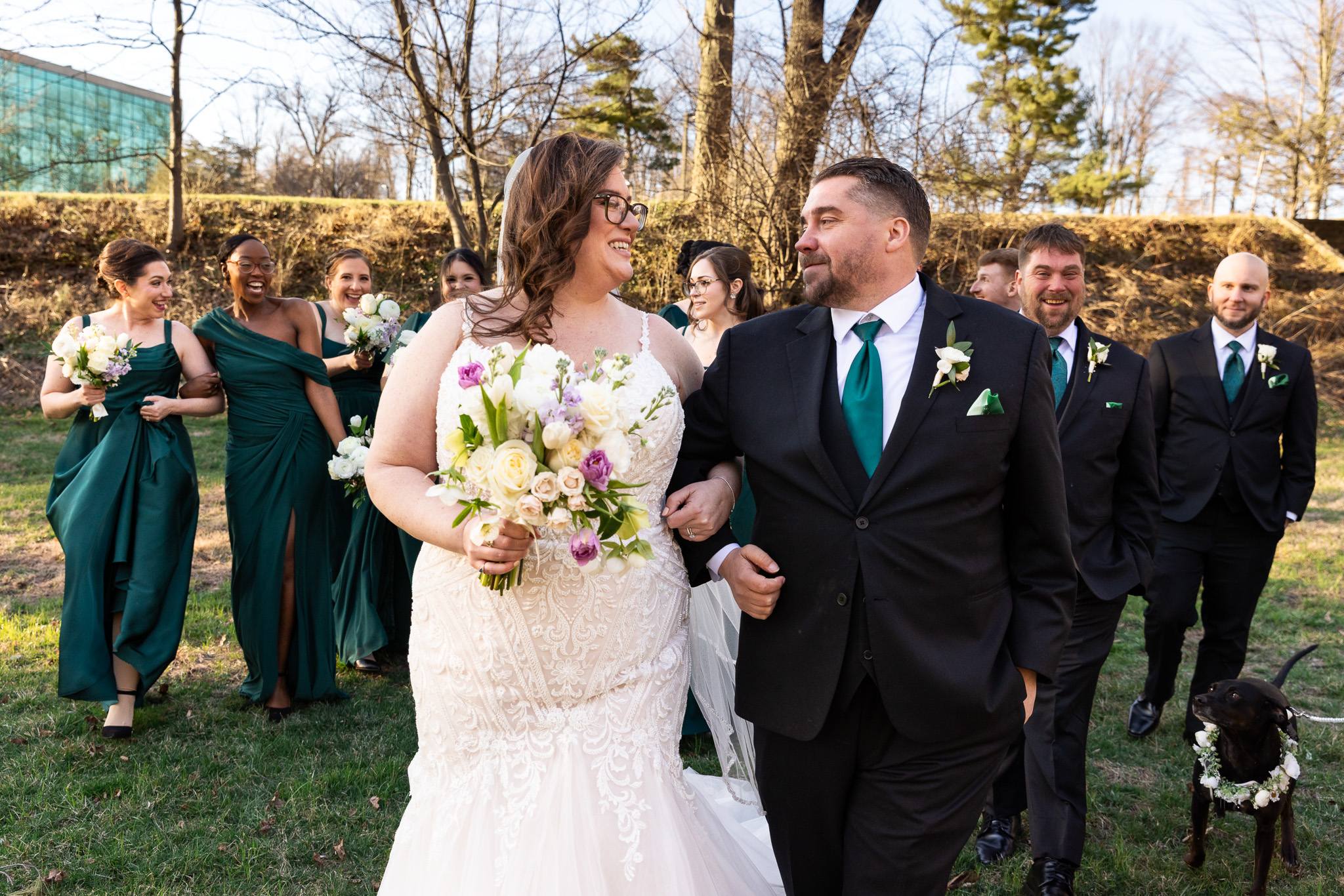 The bride and groom walk side by side after their Pomme Radnor wedding ceremony, smiling at each other as they move forward. The bride carries her bouquet, and her lace gown flows around her. Behind them, bridesmaids in deep green dresses and groomsmen in black suits follow, laughing and celebrating. A black dog wearing a floral collar walks along with the group, adding a playful detail.
