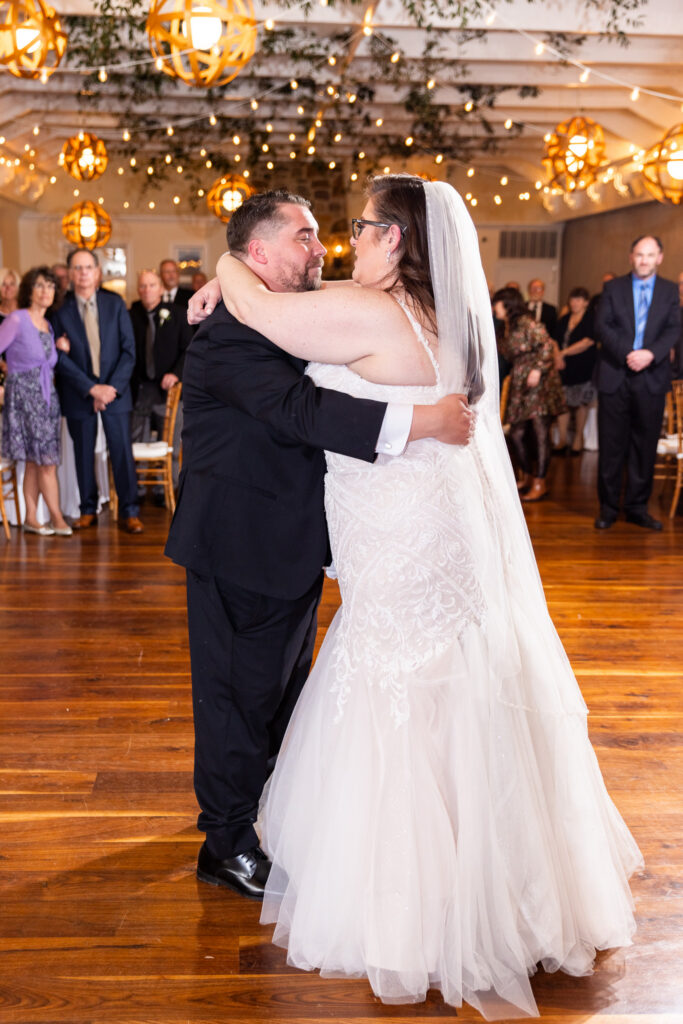 During a Pomme Radnor wedding reception, the bride and groom share their first dance in the center of a wooden dance floor. The bride’s arms rest around the groom’s shoulders while his arms wrap around her waist. Guests stand in a circle around them, watching quietly. Overhead, warm string lights and hanging fixtures create a cozy, golden glow across the room.