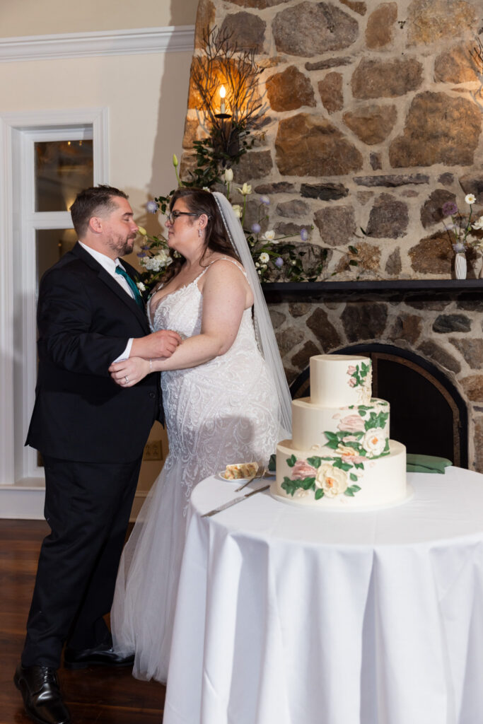 The bride and groom stand close together beside a round table holding a three-tiered wedding cake decorated with soft floral details. Behind them, a rustic stone fireplace rises up the wall, accented with greenery and a glowing candle sconce. The couple faces each other, hands gently touching, sharing a quiet, anticipatory moment before cutting the cake.