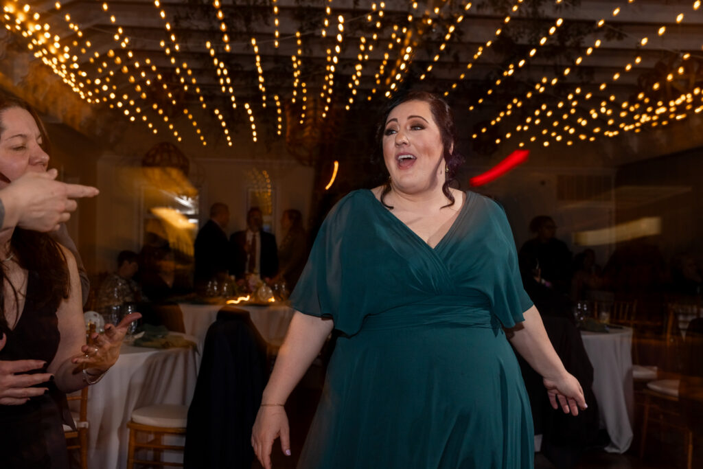 A wedding guest in a deep green, flowy dress stands on the dance floor mid-reaction, her mouth open in laughter or surprise as she gestures with her hands. Warm string lights stretch across the ceiling above her, creating a glowing canopy of golden bokeh. Other guests sit and mingle at round reception tables in the softly lit background, while motion blur from dancing and movement adds energy to the scene.