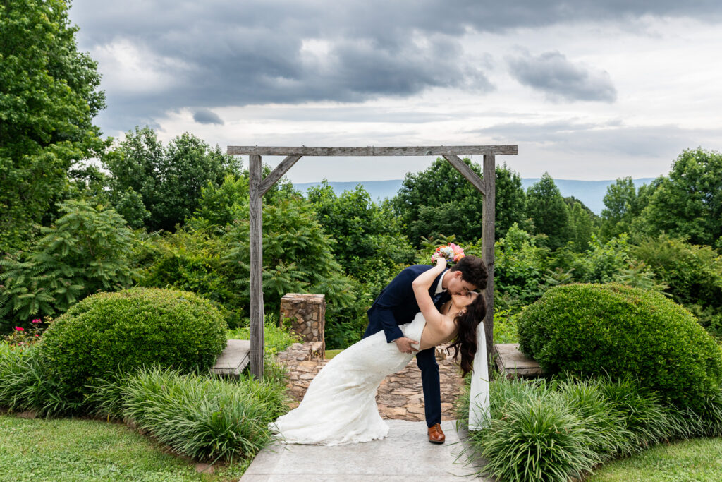 A couple shares a dramatic dip and kiss beneath a wooden arbor overlooking lush greenery and distant mountains, capturing a break in a rainy wedding day that allowed for outdoor portraits. The bride’s veil drapes toward the ground while the groom supports her in the pose.