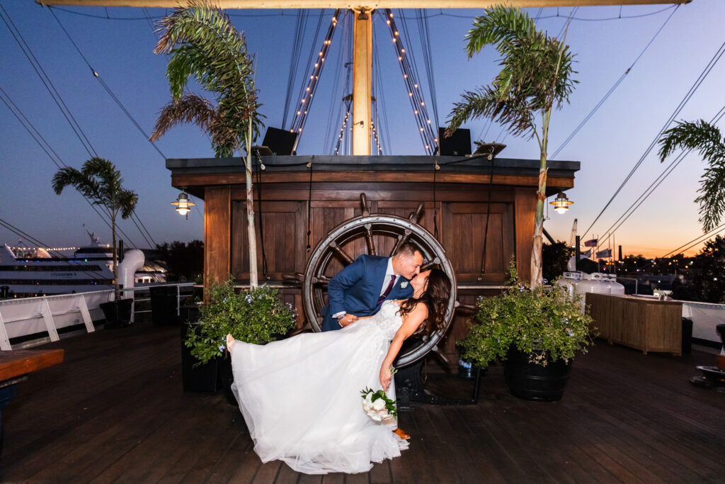 Romantic nighttime wedding portrait aboard a ship-inspired rooftop venue at sunset. The groom dips the bride backward in front of a large wooden ship wheel while string lights glow overhead and palm trees sway on either side.