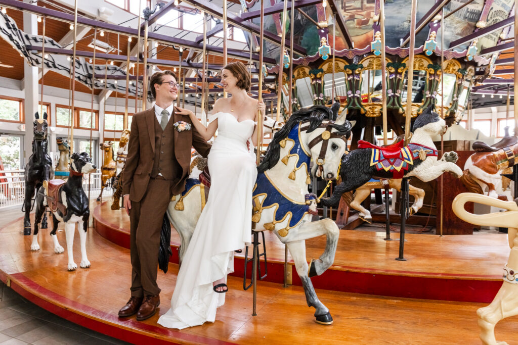 Playful wedding portrait of a bride and groom standing beside a colorful indoor carousel surrounded by painted carousel animals and glowing string lights. The bride sits sideways on a carousel horse in a modern off-the-shoulder gown while the groom stands beside her in a brown three-piece suit, both smiling warmly at each other.